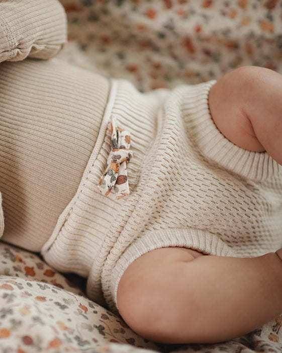 A close-up of a baby lying on a soft beige blanket, wearing a cream-colored waffle-knit romper with a small decorative animal applique at the waist.