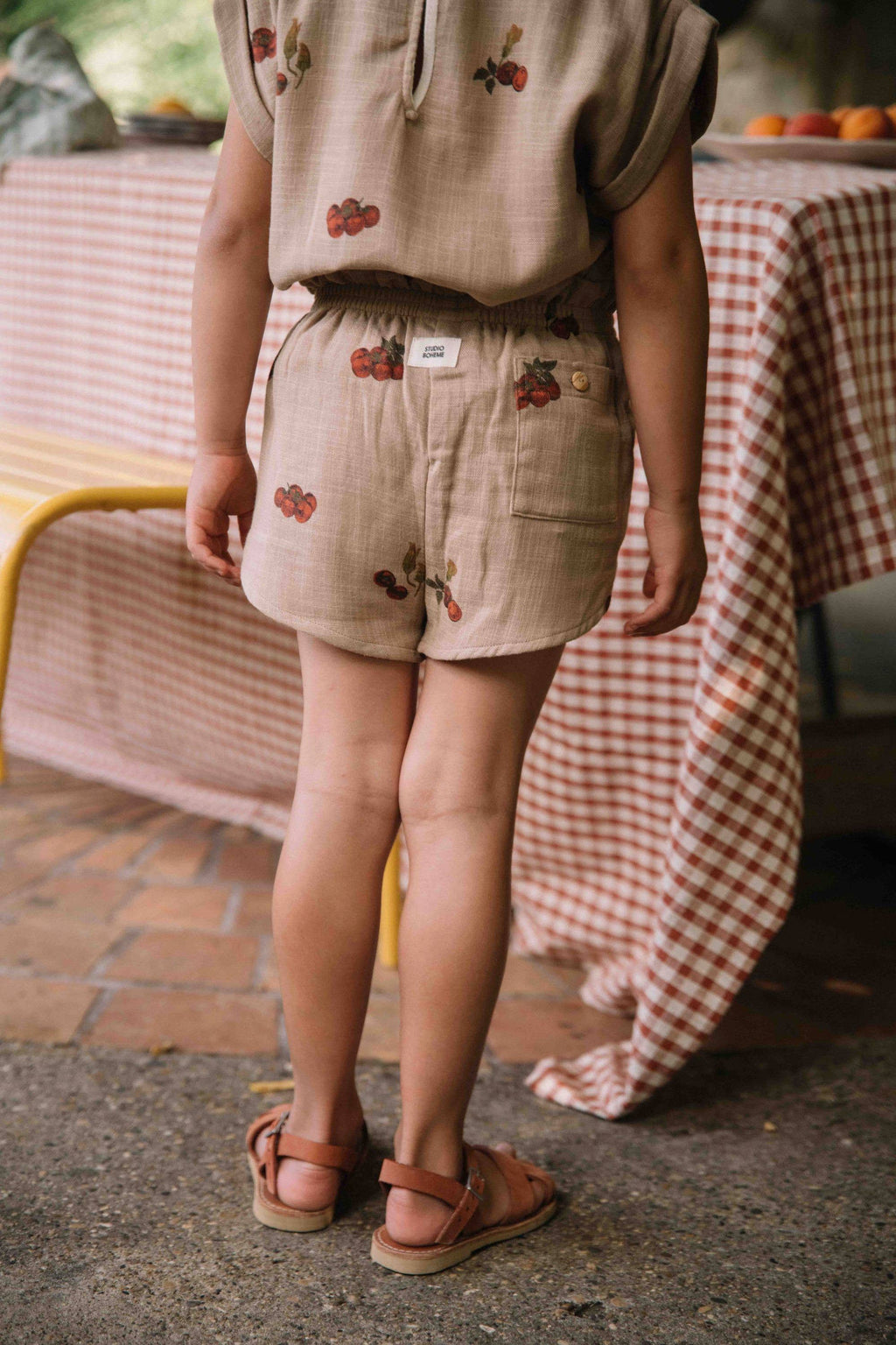 Child wearing beige shorts with floral embroidery standing outdoors near a table with a checkered tablecloth.