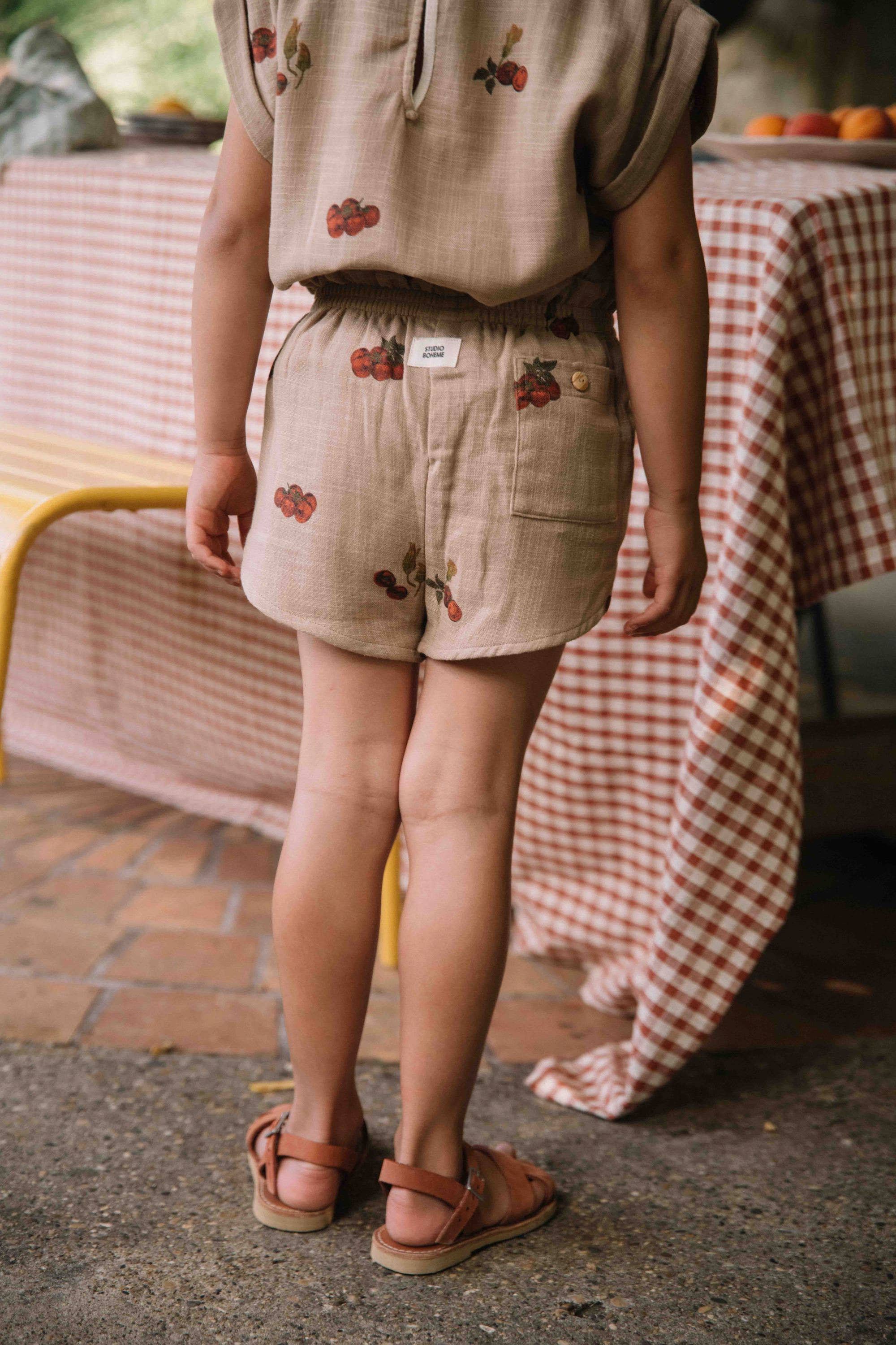 Child wearing beige shorts with floral embroidery standing outdoors near a table with a checkered tablecloth.