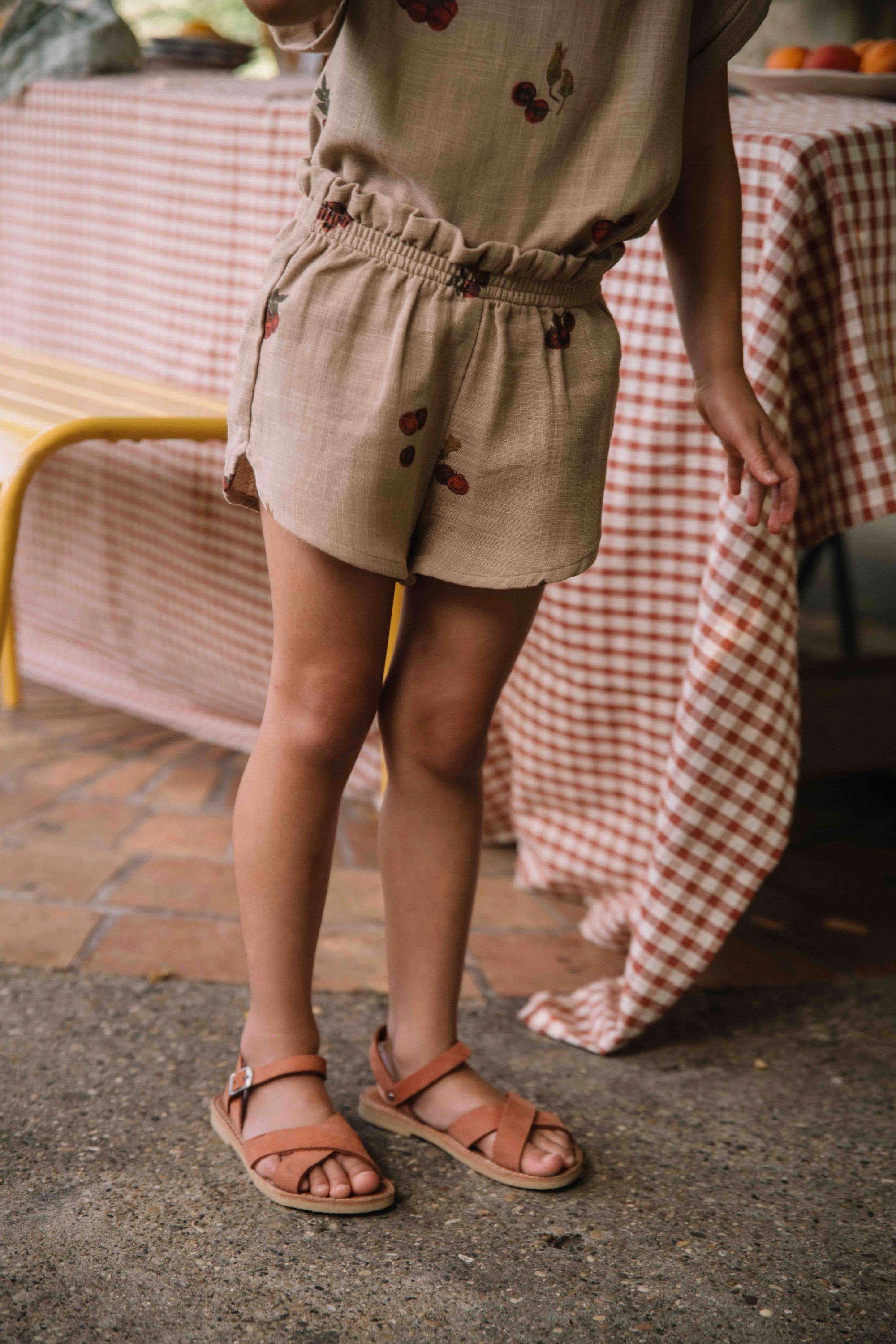 Person wearing beige shorts with cherry embroidery and brown sandals on a checkered tablecloth background.
