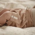 A close-up of a baby's legs and feet wearing a soft knit romper in a warm beige or light brown color, lying on a textured cream blanket.