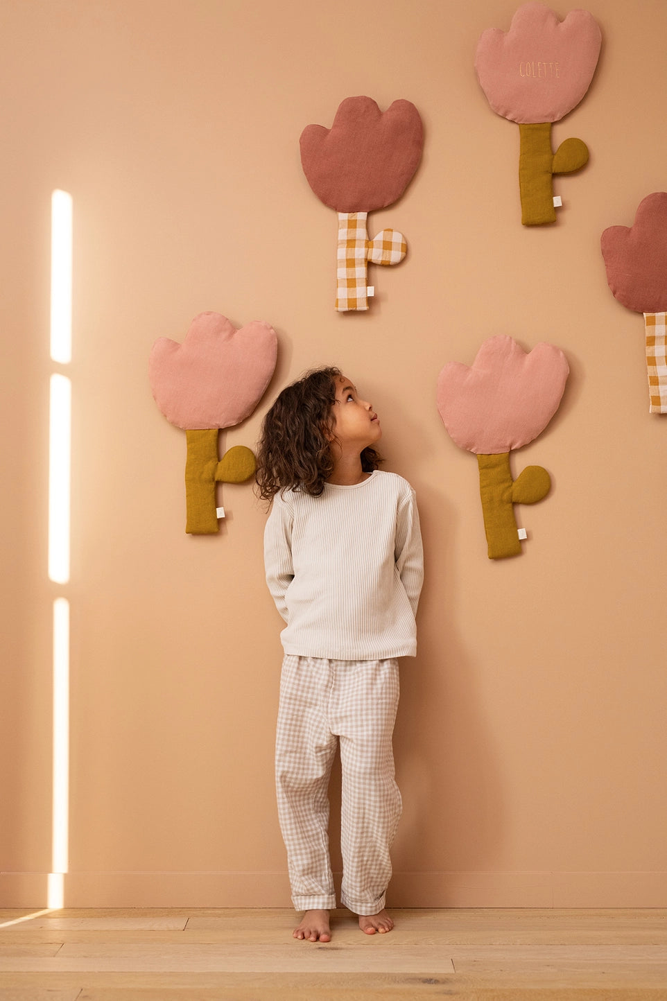 A child wearing a cream long-sleeve top and beige gingham pajama pants stands barefoot against a peach wall, looking up at large pink and brown felt flower decorations.