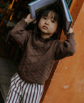 A young child wearing a brown textured knit sweater and striped pants, holding a book over their head against an orange wall.