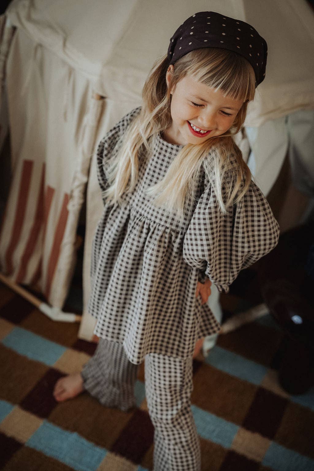 A young girl with blonde hair wearing a black and white gingham check tunic top and matching pants, along with a black polka dot headband, standing in a cozy room with a tent in the background.
