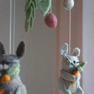 A close-up of a felt baby mobile with hanging animal figures, including a gray bunny holding an orange carrot and green leaves above, against a soft pink background.
