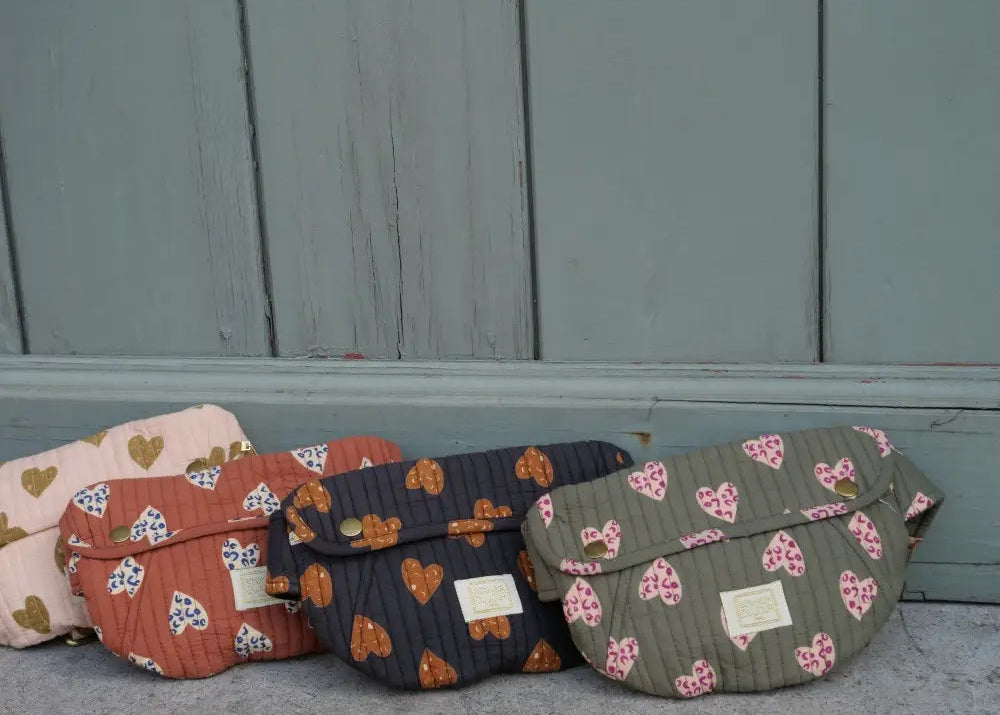 Four quilted fabric shoulder bags with floral and heart patterns lined up against a weathered blue-gray wooden wall.