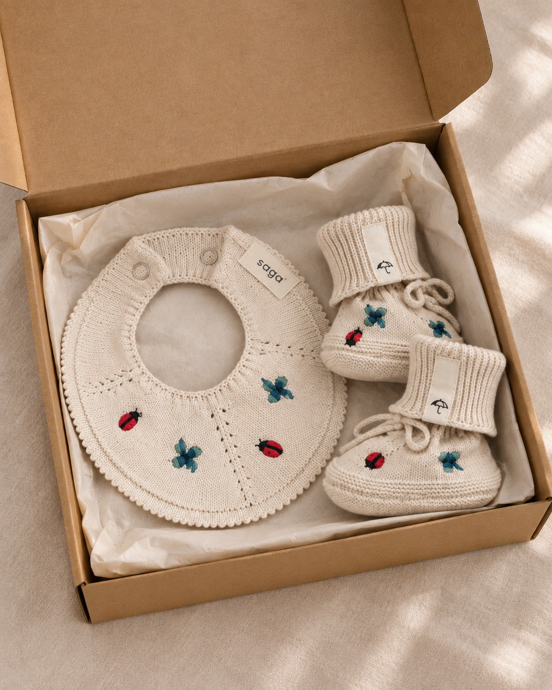 A baby gift set including a cream-colored knit bib and booties with embroidered ladybugs and butterflies, presented in an open brown cardboard box on a soft beige fabric background.