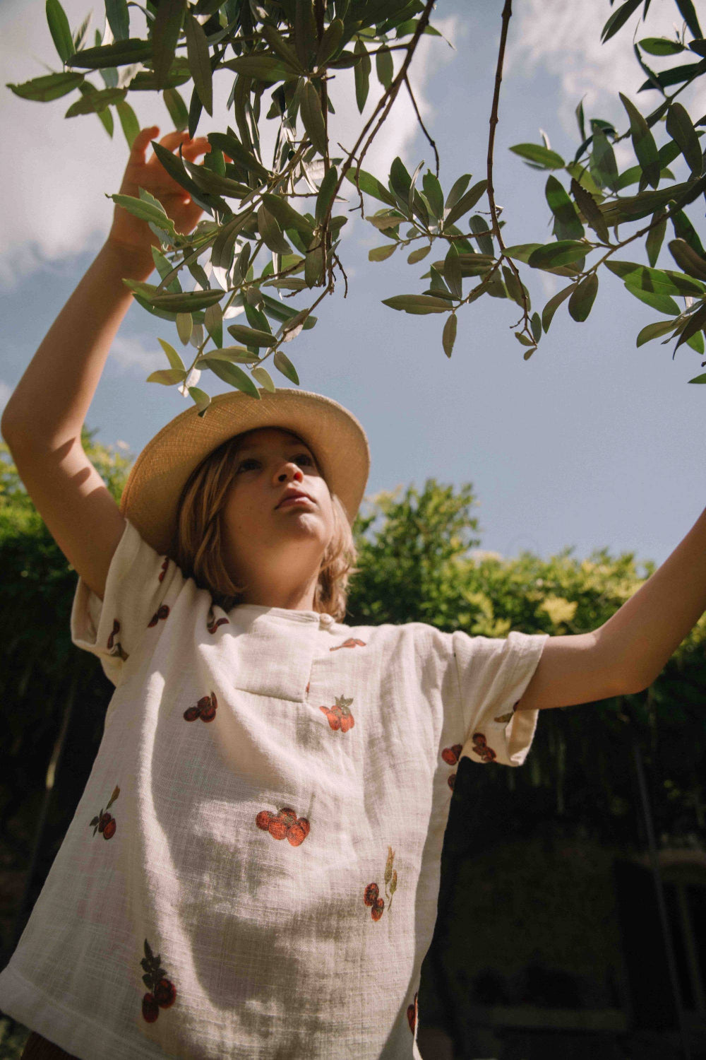 Person wearing a straw hat and white dress with cherry pattern, standing under green leaves.