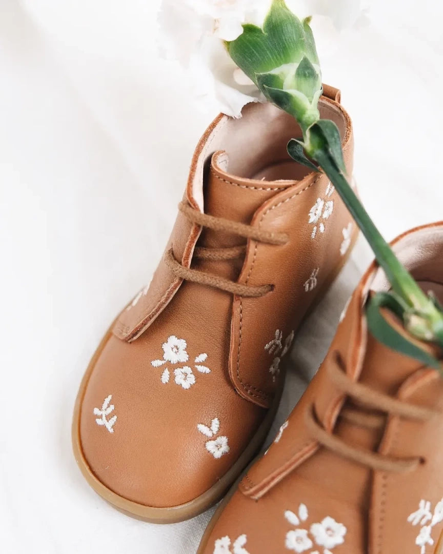 A pair of tan leather baby booties with white floral embroidery and matching laces, styled with a green flower stem on a white background.