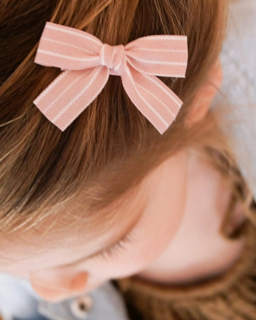 A close-up of a child with light brown hair wearing a light pink striped bow hair clip.