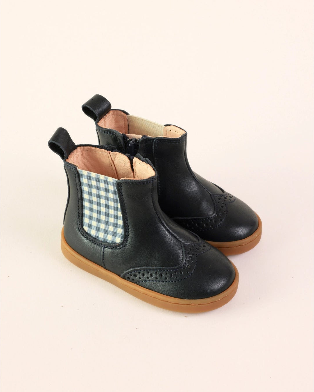 A pair of toddler black leather Chelsea boots with a blue and white gingham elastic side panel and brown rubber soles, shown on a light beige background.