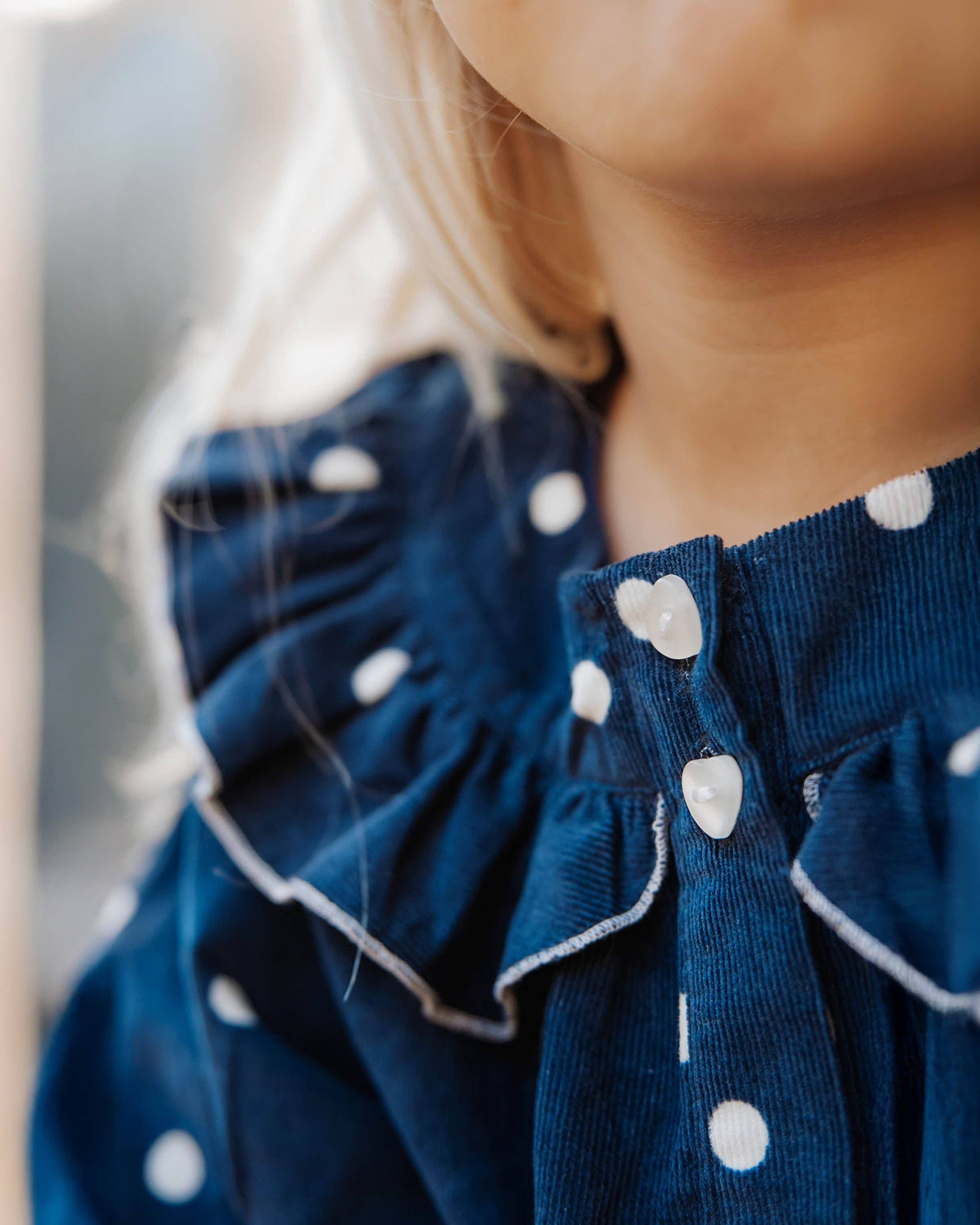 Close-up of a child wearing a navy blue corduroy top with white polka dots and ruffled trim, featuring decorative white buttons at the collar.