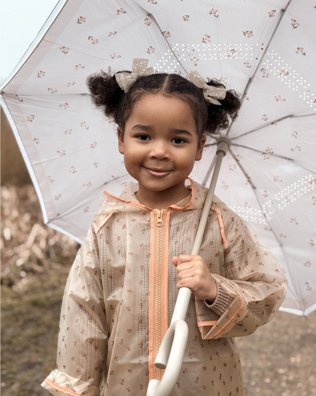 Child holding a transparent umbrella with floral patterns, wearing a beige raincoat.