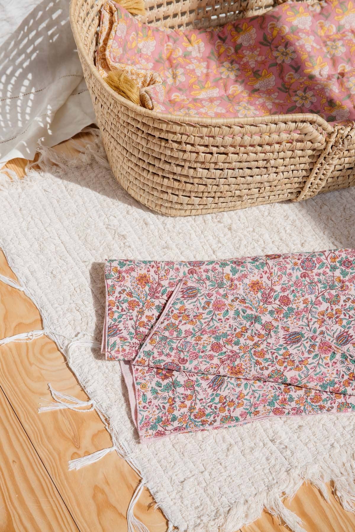 A folded piece of pink floral fabric with a small-scale botanical print is displayed on a white textured rug next to a woven basket containing matching pink floral fabric.