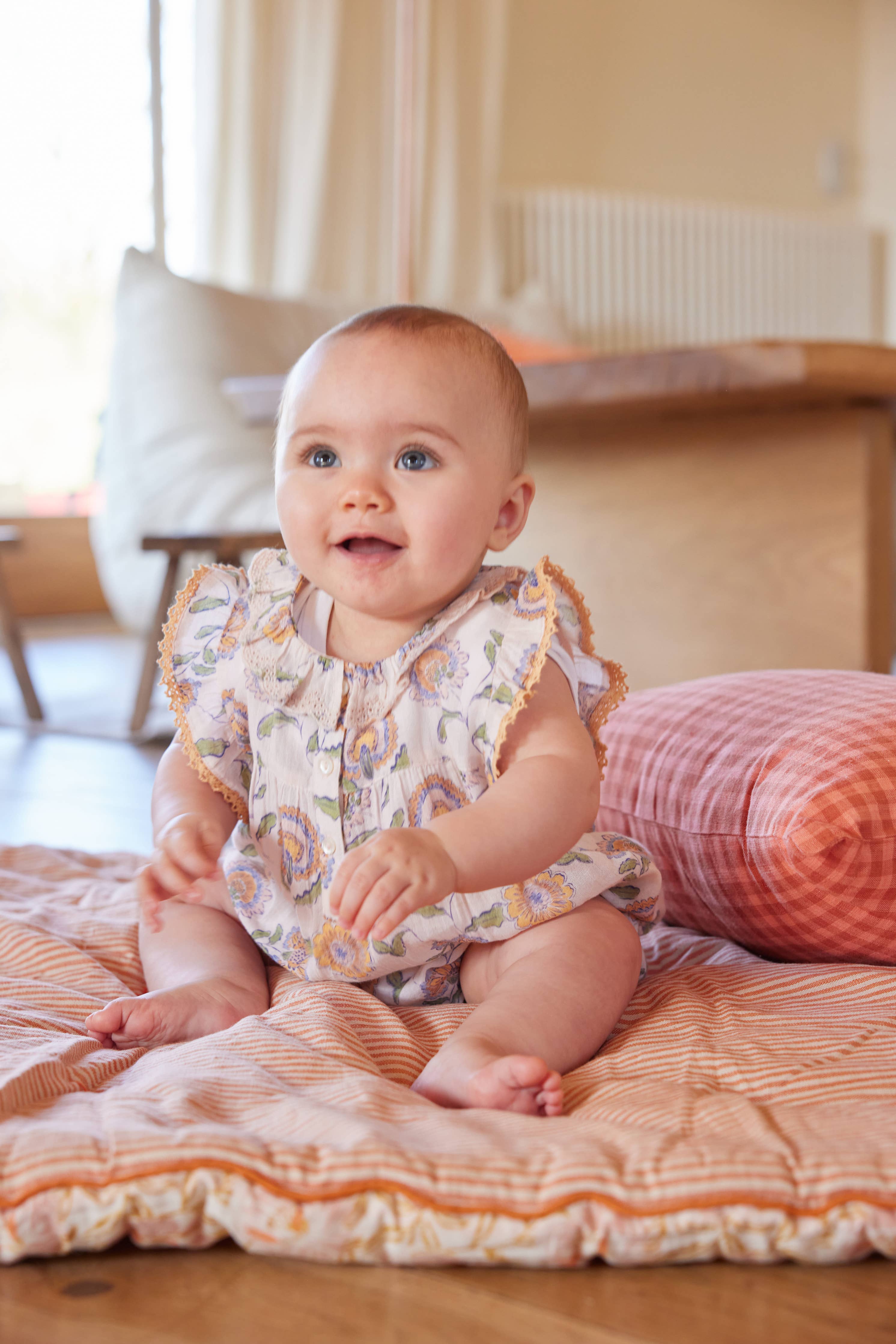 Baby sitting on a bed wearing a floral outfit with ruffled sleeves.