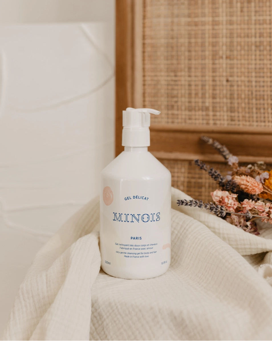 A white pump bottle of Minois ParisGel Delicat body wash resting on a textured cream linen cloth, styled with dried flowers against a background of a woven cane panel and white ceramic tile.