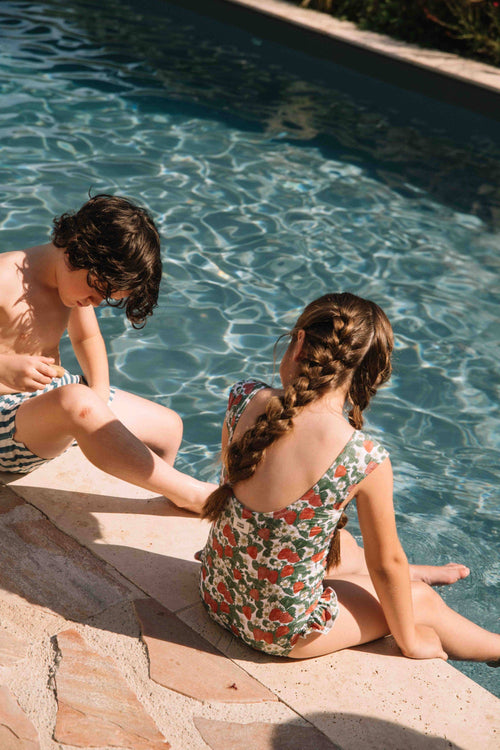 Two children sitting by a pool, one applying sunscreen to the other.