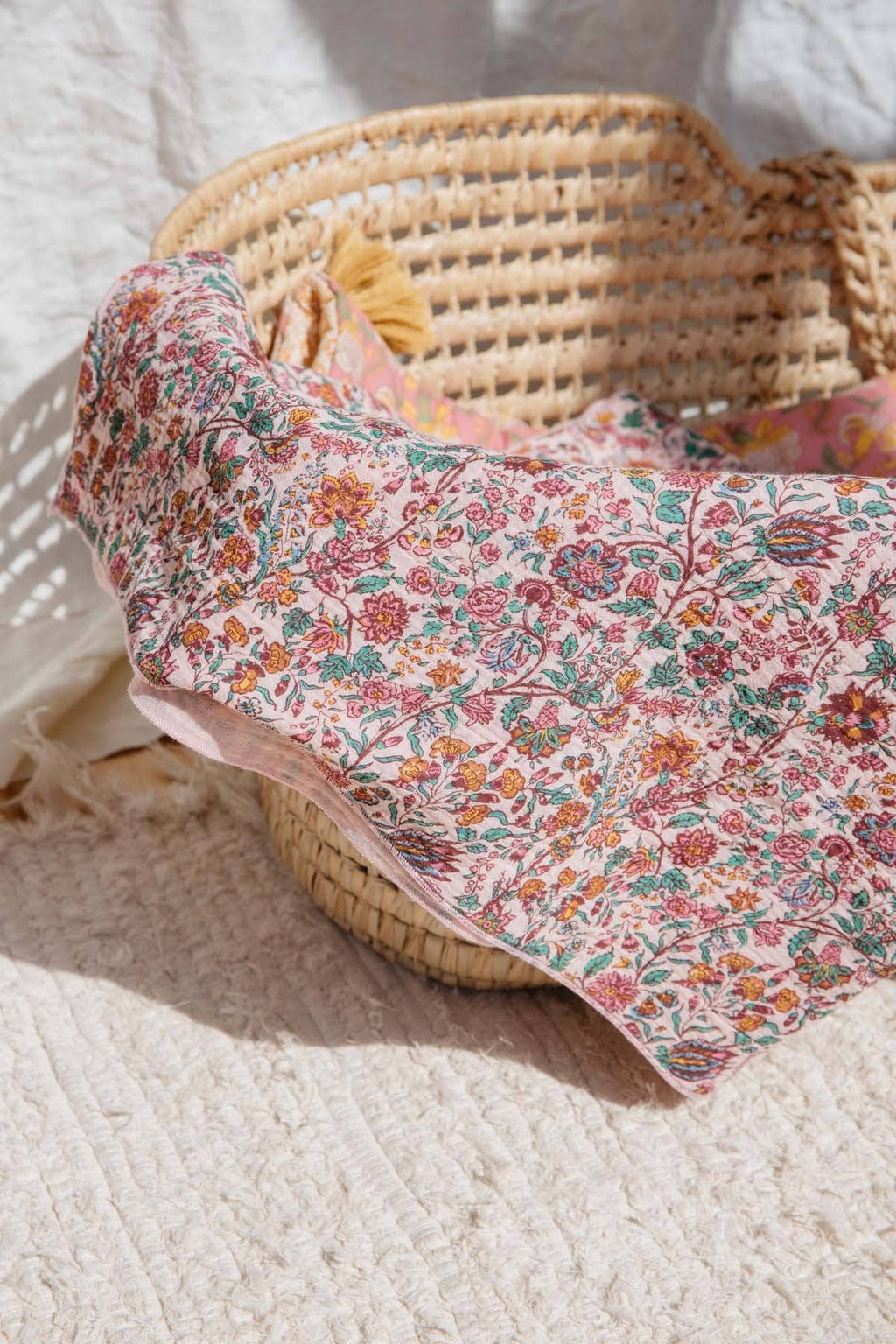 A floral baby blanket with a pink, orange, and green pattern draped over a woven straw basket on a textured cream rug.