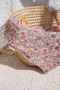 A floral baby blanket with a pink, orange, and green pattern draped over a woven straw basket on a textured cream rug.