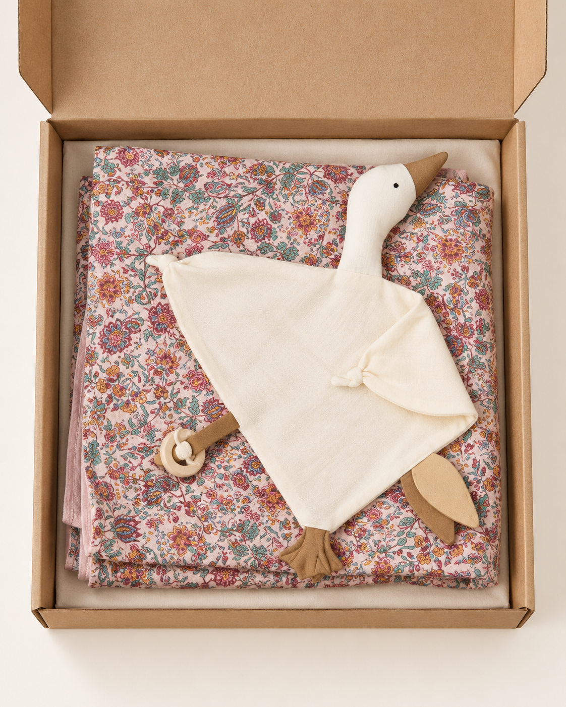 A baby comforter set in a kraft gift box, featuring a cream-colored goose-shaped security blanket with a wooden teething ring and a folded pink, red, and blue floral patterned blanket underneath.