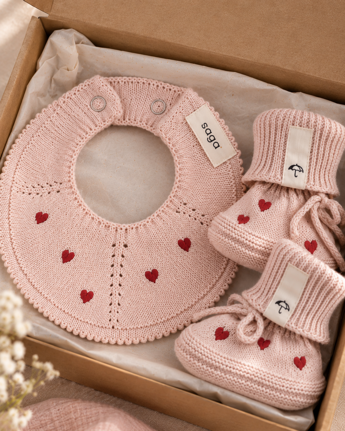 A baby gift set in a brown cardboard box, featuring a light pink knitted bib and booties with small red embroidered hearts. The brand tag 'caga' is visible on both items.