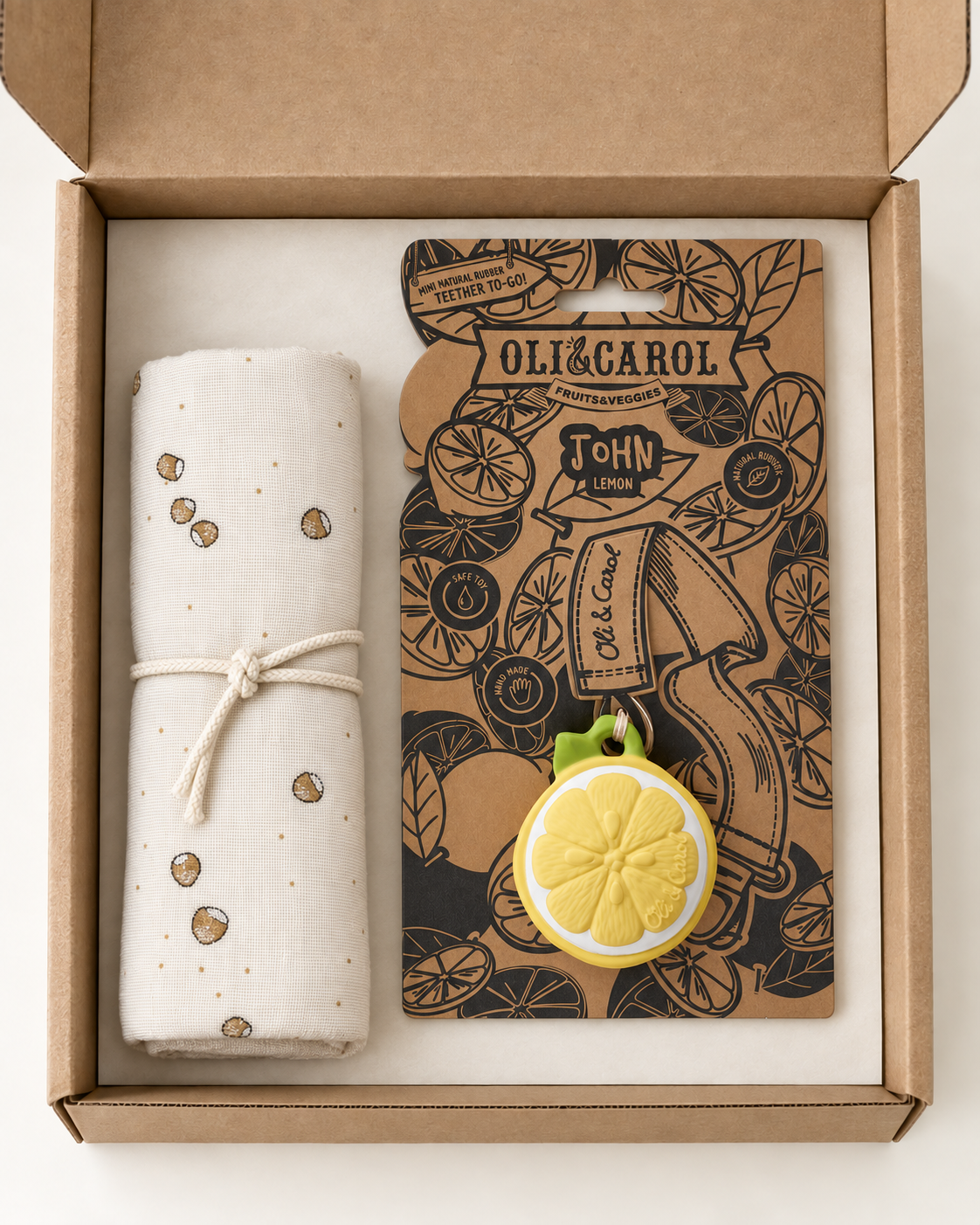 An open kraft gift box containing a cream-colored rolled fabric item with small brown leaf prints tied with a string, next to a branded card for Oli & Carol John Lime. The card features black botanical illustrations and a yellow lemon-shaped charm with green loops at the top.