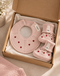 A baby gift set in a kraft paper box, featuring a light pink knitted bib with red heart details and a matching pair of booties, both with white brand tags reading 'sofa'.