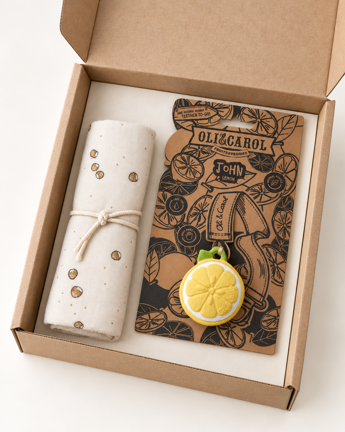 An open kraft paper gift box containing a rolled cream fabric item tied with a string and a brown illustrated card or notebook with lemon-themed artwork and the visible brand name OligCarol.
