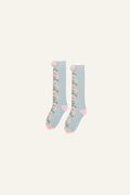 A pair of light blue socks with a pink and green floral pattern down the side, featuring pink heels and toes, and pom-poms at the top, shown on a light gray background.