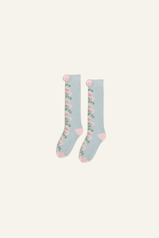 A pair of light blue socks with a pink and green floral pattern down the side, featuring pink heels and toes, and pom-poms at the top, shown on a light gray background.