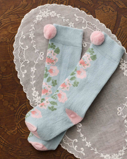 A pair of light blue knee-high socks with a pink and green floral pattern, featuring pink pom-poms at the top and pink heels, displayed on a white lace doily over a dark brown patterned surface.