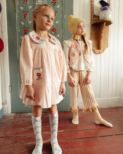 Two young girls modeling vintage-style clothing in a rustic room. The girl in the foreground wears a pale pink long-sleeve dress with a floral Peter Pan collar, matching floral knee-high socks, and a pink bonnet.