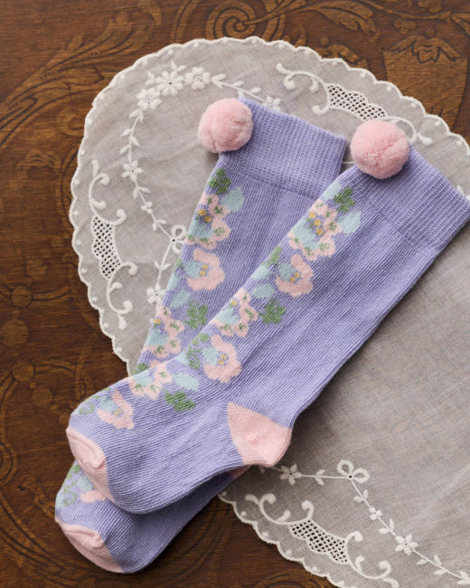 A pair of lavender knee-high socks with a pastel floral pattern and pink pom-poms at the top, displayed on a white lace doily over a dark wood background.