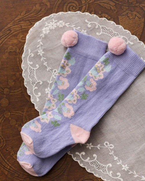 A pair of lavender knee-high socks with a pastel floral pattern and pink pom-poms at the top, displayed on a white lace doily over a dark wood background.