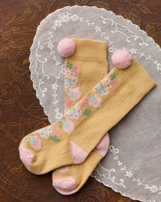 A pair of mustard yellow crew socks with a pastel floral pattern and pink pom-poms on the tops, displayed on a white lace doily over a dark brown wooden surface.