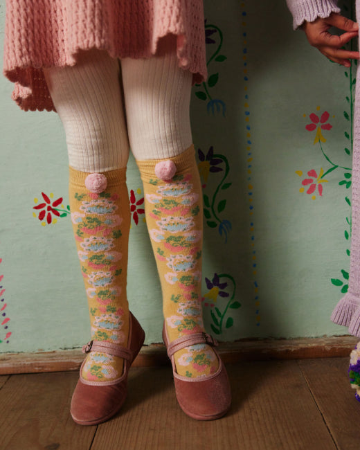 A person wearing pastel pink and yellow floral knee-high socks with pom-pom details, paired with a pink skirt and pink shoes, standing against a green floral wall.