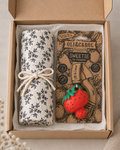 A gift box containing a rolled cream fabric item with a blue floral pattern tied with a white string, next to a branded card for OLI&CAROL Sweetie the Strawberry. The card features a 3D strawberry charm.