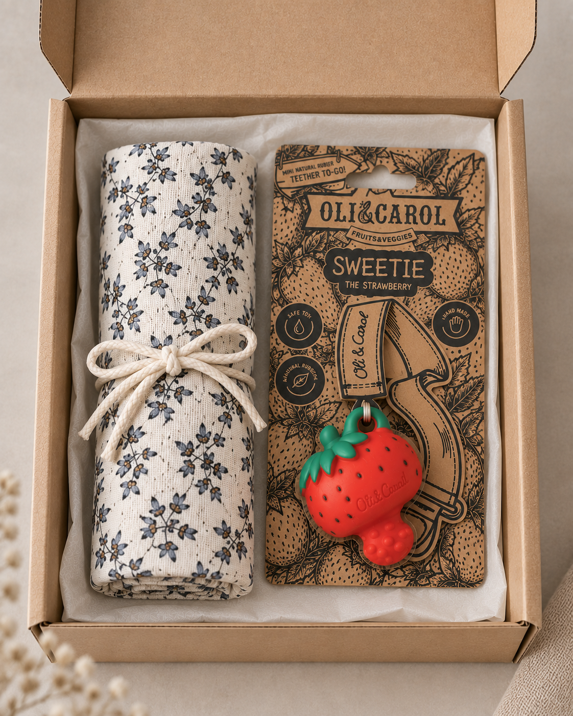 A gift box containing a rolled cream fabric item with a blue floral pattern tied with a white string, next to a branded card for OLI&CAROL Sweetie the Strawberry. The card features a 3D strawberry charm.