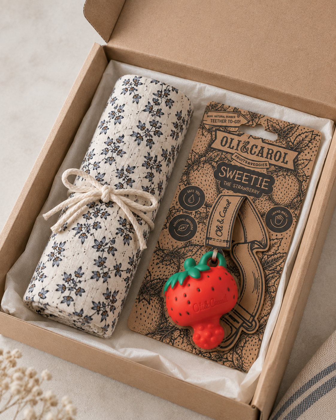 A gift box containing a rolled cream fabric item with a dark floral print tied with a string, next to a card for an Oli&Carol Sweetie teether. The teether is a red strawberry-shaped rubber toy with green leaves.