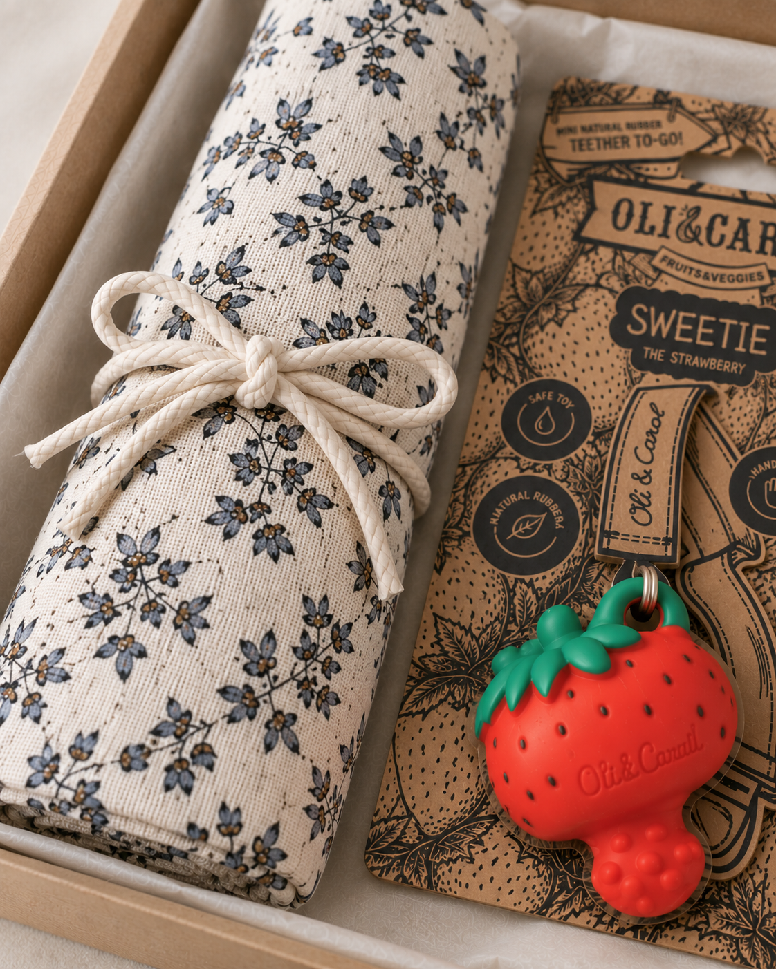 A gift set from Old Cannibals featuring a rolled cream fabric item with a blue floral print tied with a white cord, next to a brown paper bag labeled 'Oli & Carol Sweetie the Strawberry' and a small red strawberry-shaped charm.